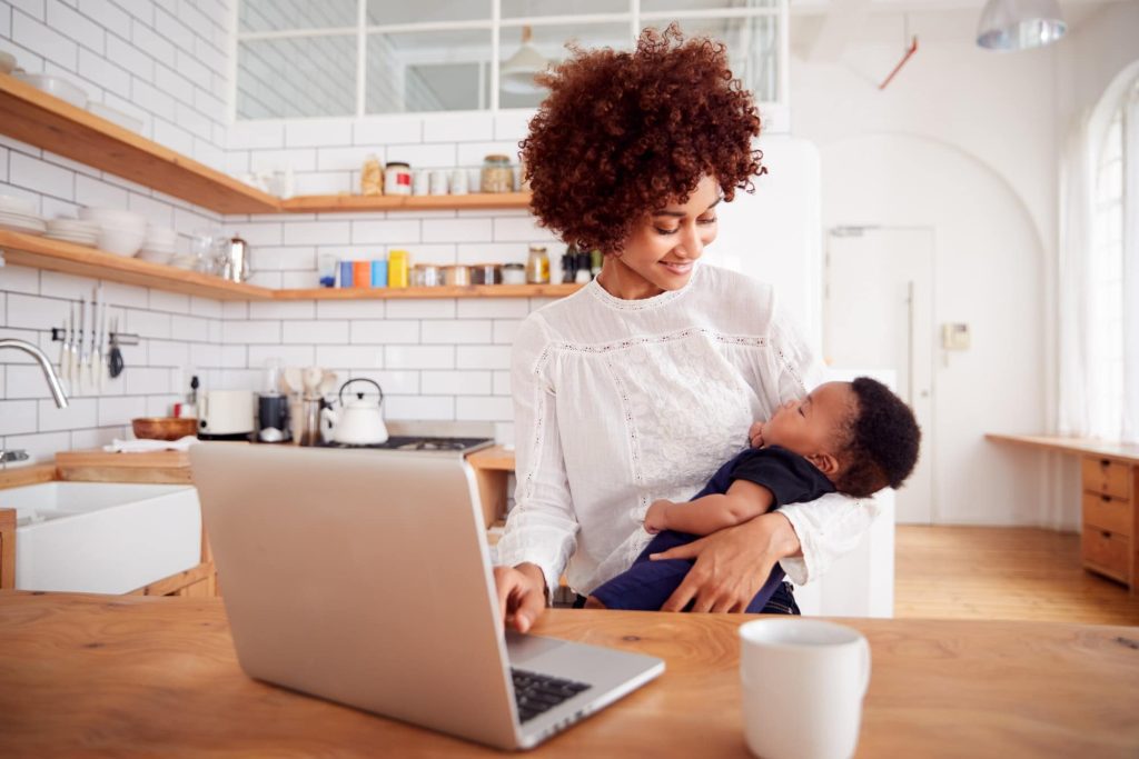 Multi-Tasking Mother Holds Sleeping Baby Son And Works On Laptop Computer In Kitchen