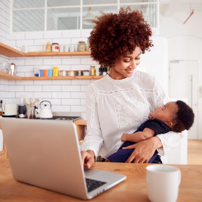 Multi-Tasking Mother Holds Sleeping Baby Son And Works On Laptop Computer In Kitchen Multi-Tasking Mother Holds Sleeping Baby Son And Works On Laptop Computer In Kitchen